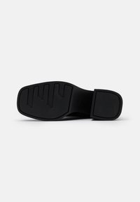 Black shoe sole with a textured surface, featuring a block heel and a segmented tread pattern for grip. Smooth edges and minimal branding.