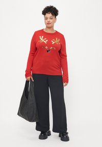 Pulôver vermelho com aplicações de rena em lantejoulas douradas, combinado com calças largas pretas. A modelo segura uma mala de tote de couro preto e usa sapatos chunky.