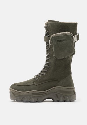 Wysokie buty w kolorze oliwkowej zieleni wykonane z zamszu, z grubą podeszwą z protektorem, sznurowane z przodu oraz posiadające boczną kieszonkę na zamek, przymocowaną paskiem z klamrą.