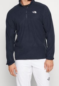 Marinblå fleece-pullover med en kvart dragkedja och ribbad krage, med en logotyp på vänster bröst och långa ärmar.