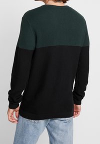 Pull en tricot avec une partie supérieure texturée verte et une partie inférieure noire unie. Manches longues avec des poignets et un ourlet côtelés.