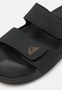Sandale noire à glissière avec deux sangles réglables en velcro, surface texturée et accent de logo brun. Fabriquée à partir de matériaux doux et durables.