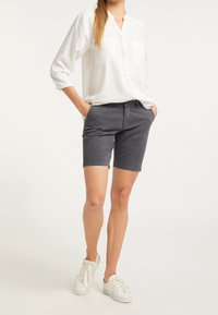 Donna che indossa una camicia bianca a maniche lunghe, pantaloni corti grigio scuro fino al ginocchio, sneakers bianche, in piedi con le mani in tasca su uno sfondo neutro.