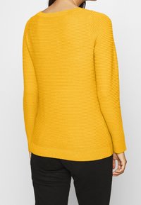 Pull en maille côtelée jaune avec un col rond et des manches longues, présentant une texture subtile et un ourlet inférieur ajusté.