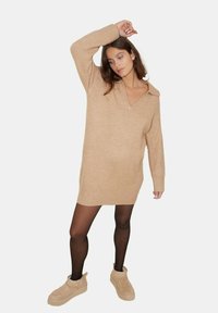 Robe-pull beige en maille avec un col en V et des manches longues, associée à des collants noirs et des bottines beige à la finition douce et texturée.