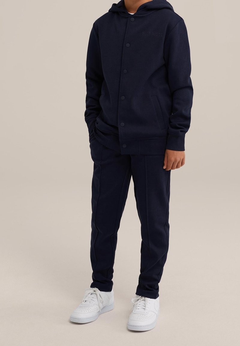 Dziecko ubrane w granatową bluzę z kapturem i dopasowane spodnie, w białe trampki, stojące na tle jednolitego beżowego tła.