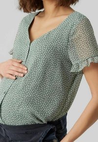 Grüne Bluse mit weißen Punkten, V-Ausschnitt und kurzen, gerafften Ärmeln, aus leichtem Stoff, mit durchgehender Knopfleiste vorne.