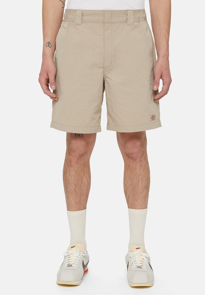 Shorts beiges en tissu léger, avec un devant plat, des poches latérales et un logo brodé sur la jambe gauche.