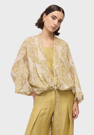 Mujer con una chaqueta beige holgada con patrones dorados de mariposas sobre una camiseta amarilla y pantalones de pierna ancha a juego, posando frente a un fondo liso.