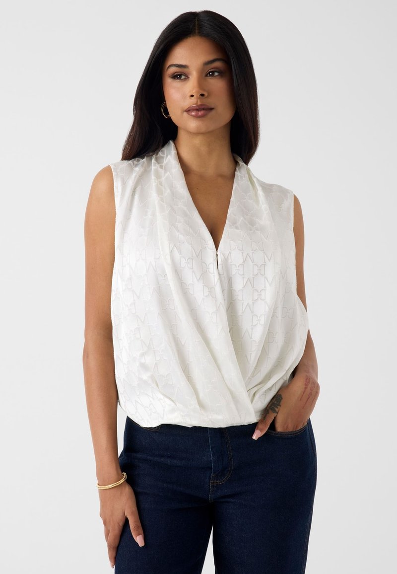 Femme aux longs cheveux foncés porte un chemisier blanc sans manches à nouer et un jean bleu foncé, debout avec une main dans la poche devant un fond uni.