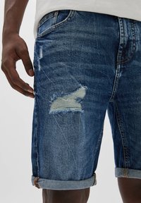 Džínové šortky ve tmavě modré barvě s opotřebovaným vzhledem, s viditelnými dírami a ohrnutým lemem. Texturovaná tkanina s mimovanými švy.