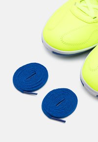Zapatillas deportivas de color amarillo neón con un upper texturizado, acompañadas de dos cordones redondos de color azul oscuro sobre un fondo blanco.