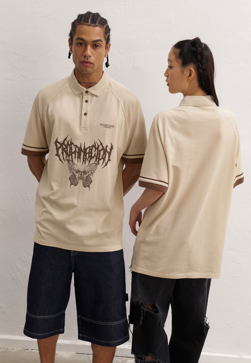 Beige pikéskjorte med grafisk design av en sommerfugl og hodeskalle. Brune striper ved kragen og ermene. Kombinert med mørke denimshorts.
