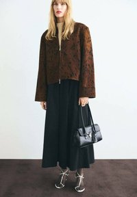 Veste en fausse fourrure marron tachetée avec une fermeture éclair, associée à une longue jupe noire et des talons transparents à lanières, tenant un sac à main noir.