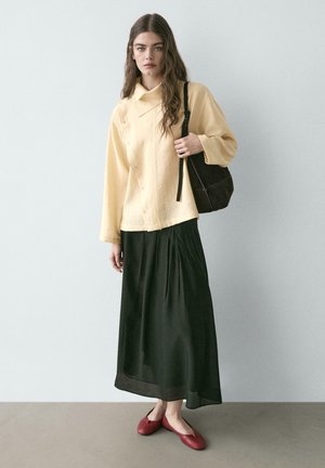 Frau trägt ein locker sitzendes, gelbes Oberteil mit Knöpfen, einen langen schwarzen Rock, rote Ballerinas und hält eine schwarze Umhängetasche vor einer schlichten Wand.