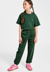 Grünes Baumwoll-T-Shirt mit einer Brusttasche und passenden grünen Jogginghosen. Das Outfit wird mit rosa Sneakern mit weißen Akzenten kombiniert.