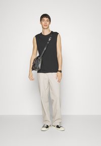 Maglietta nera senza maniche, pantaloni ampi di colore chiaro e sneakers in bianco e nero. Borsa a tracolla nera con due tasche. Texture liscia.