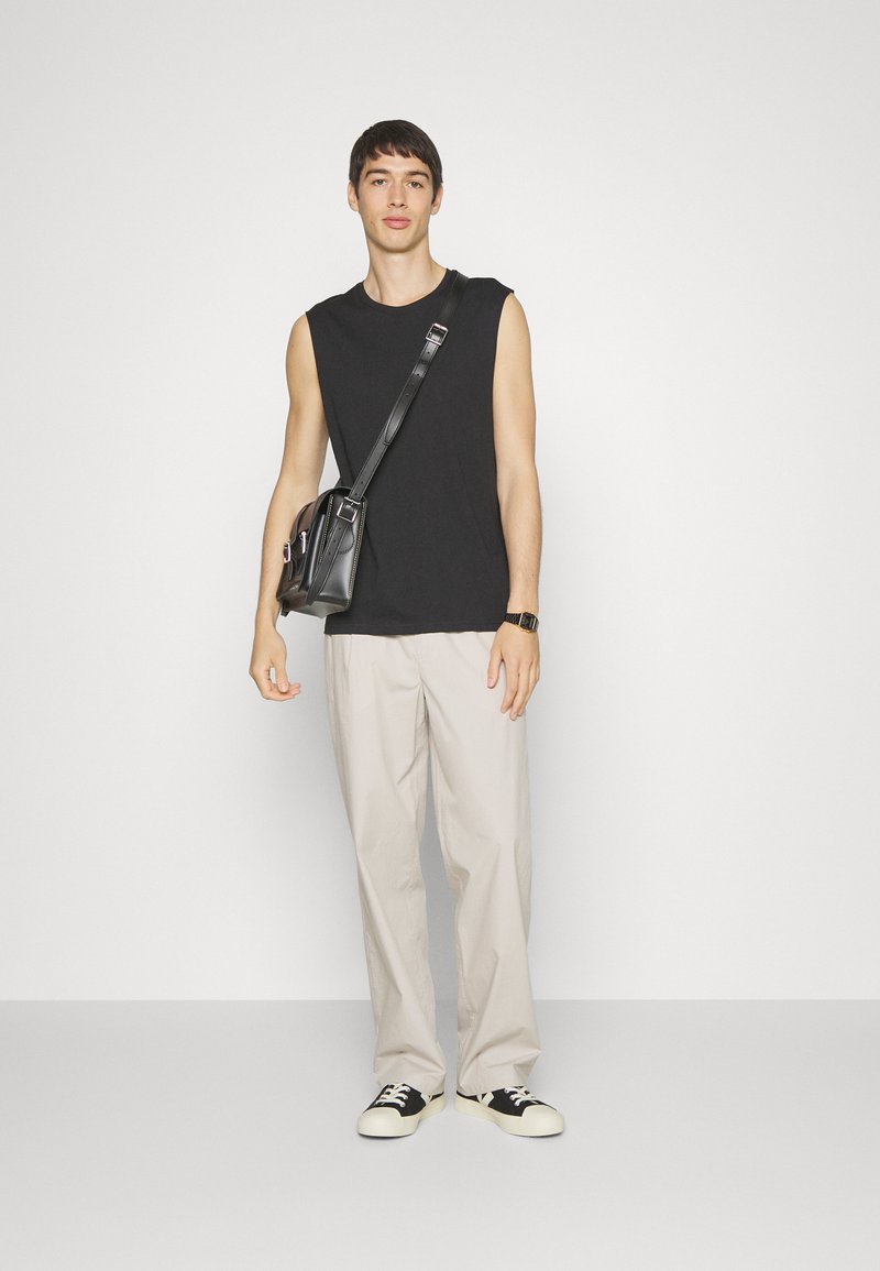 Maglietta nera senza maniche, pantaloni ampi di colore chiaro e sneakers in bianco e nero. Borsa a tracolla nera con due tasche. Texture liscia.
