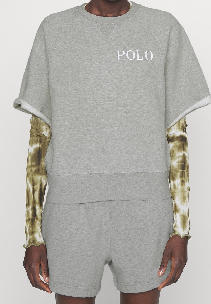 Persoon draagt lichtgrijze korte broek en een grijze sweatshirt met korte mouwen met de tekst "POLO", gecombineerd met een lange mouwen tie-dye shirt eronder.