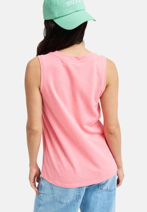 Kobieta ubrana w różowy bezrękawnik, niebieskie szorty dżinsowe i zieloną czapkę z daszkiem z napisem „ROXY”, widziana od tyłu.