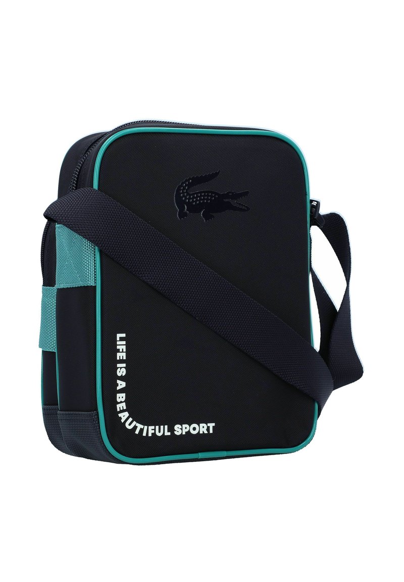 Sac à bandoulière en tissu noir avec des accents turquoise, présentant un logo crocodile texturé et l'inscription "LA VIE EST UN SPORT MAGNIFIQUE" en blanc.