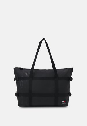 Bolso tote rectangular negro con asas largas, correas con hebilla a los lados y parche pequeño con el logo de la marca en la esquina inferior derecha.