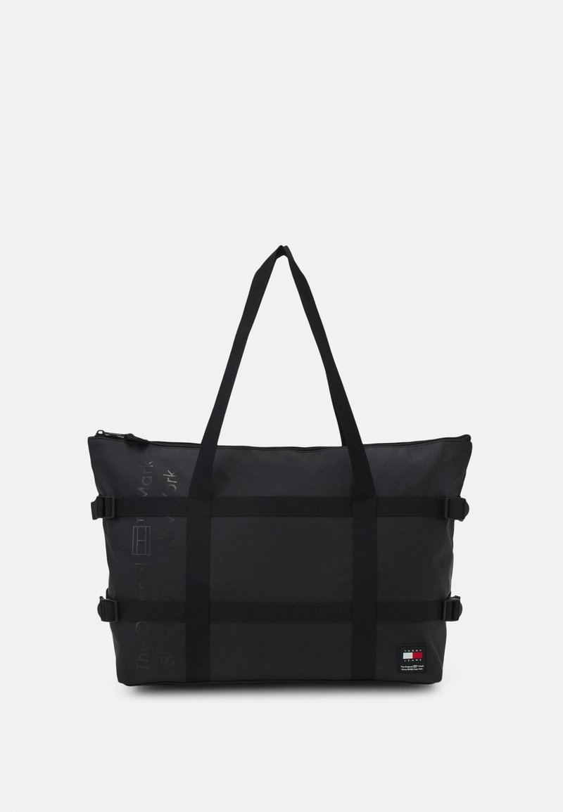 Sac fourre-tout rectangulaire noir avec longues anses, sangles à boucle sur les côtés et petit logo de la marque en bas à droite.