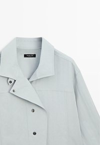 Giacca grigio chiaro con colletto ampio, caratterizzata da una chiusura a doppio bottone, tessuto opaco e dettagli di cuciture minimaliste.