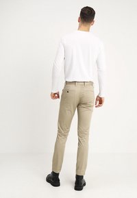 Béžové úzké kalhoty z lehké látky, s kapsami na zadní straně a poutky na pásek. Model nosí bílou košili s dlouhým rukávem a černé boty.