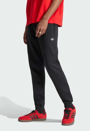 Persoon draagt zwarte Adidas joggers met wit logo, rode Adidas sneakers met zwarte strepen en een rood shirt, staand tegen een effen achtergrond.