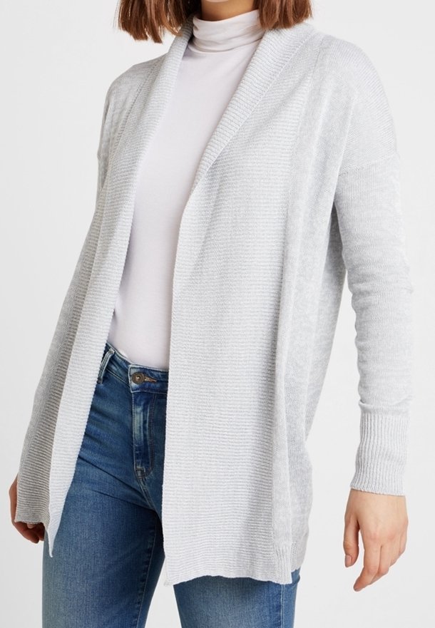 Femme portant un cardigan gris clair ouvert sur un col roulé blanc et un jean bleu, visible du cou aux hanches.
