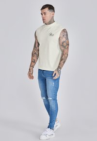 Top sin mangas beige con escote curvado, combinado con jeans ajustados azules rasgados y zapatillas blancas con acentos grises.