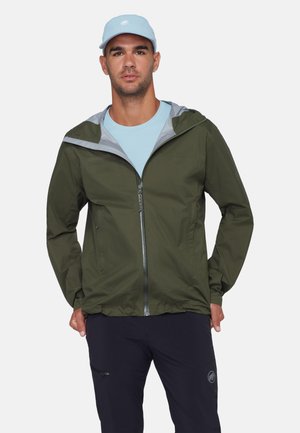 Mann trägt eine olivgrüne Jacke mit Reißverschluss und Kapuze, hellblaues T-Shirt, dunkelblaue Hose und hellblaue Kappe, steht mit den Händen in den Taschen.