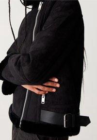 Chaqueta de ante sintético negra con cierre de cremallera, acabado texturizado y puños de piel suave. Presenta una correa de cuero en la cintura para ajustar.