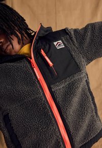 Giacca in fleece e nylon con corpo grigio e maniche nere, zip arancione brillante e patch con logo sul lato sinistro del petto.