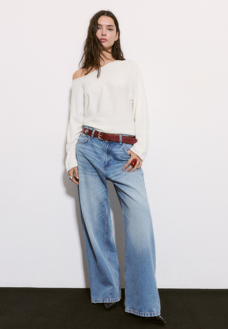 Svetlý biely pletený sveter s odhalenými ramenami, svetlomodré široké džínsy, hnedý opasok a čierne špicaté topánky. Hladká textúra s ležérnym strihom.