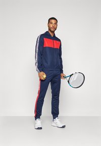 Marineblå træningsdragt med røde paneler, lynlåslukning og logo detaljer. Slim-fit bukser med sidelommer. Hvide sportssko og tennisracket inkluderet.