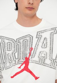 Valkoinen puuvillateeppä, jossa on iso musta "JORDAN" -kirjoitus ja punainen siluetti hyppäävästä pelaajasta, korostaen rohkeaa graafista muotoilua.