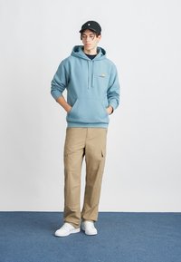 Lys blå hettegenser med frontlommer og logo, beige cargobukser med sidelommer, og hvite sneakers, iført en svart cap.