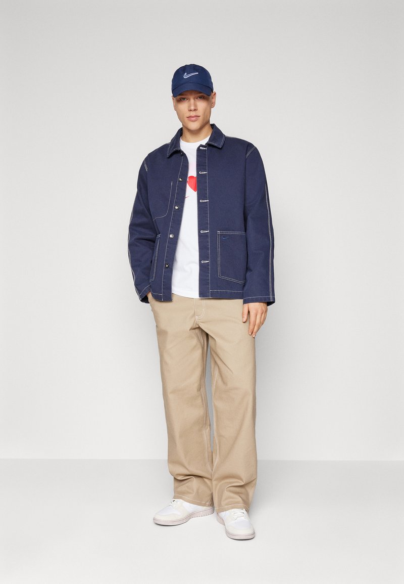 Chaqueta de mezclilla azul marino con costuras beige, camiseta blanca con un corazón rojo, pantalones beige y zapatillas blancas complementadas con una gorra azul marino.