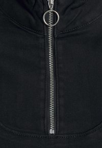 Černá mikina na zip s kovovým zipem stříbrné barvy, která má kulaté stahovací očko a viditelné švy podél švů.