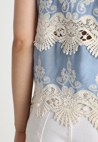 Niebieski top z tkaniny, ozdobiony skomplikowanym białym koronkowym wykończeniem wzdłuż brzegów i ramion, z kwiecistymi wzorami oraz falbaniastymi krawędziami.