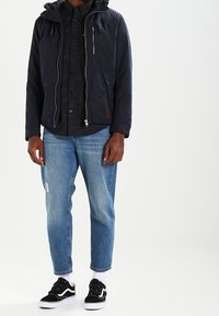 Marinblå hoodie-jacka med dragkedja fram, ihop med en mörk skjorta med knappar, ljusblå jeans och svarta sneakers med vita detaljer.