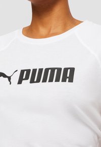 Valkoinen puuvillapaita, jossa on musta "PUMA"-logo edessä, pyöreä pääntie ja teksturoitu olkapäiden yksityiskohta.