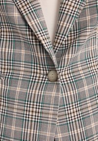 Marynarka w kratę z mieszanką zielonych, czarnych i beżowych wzorów. Zauważalne okrągłe zapięcie na guzik oraz gładki, teksturowany materiał.