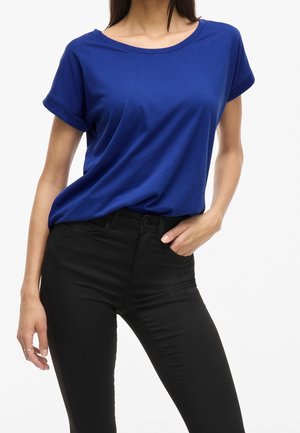 Kvinde iført en kongeblå kortærmet t-shirt, stoppet ned i sorte højttaljede skinny jeans med den ene hånd i lommen.