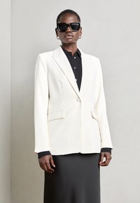 Weißes Blazer mit strukturierten Schultern, Ein-Knopf-Verschluss, zwei Vordertaschen und schwarzem Hemd darunter. Model trägt übergroße Sonnenbrille.
