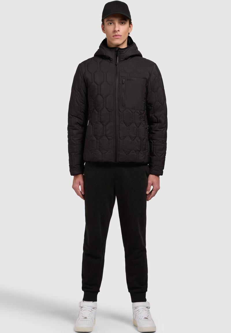 Veste rembourrée noire avec motif hexagonal, fermeture éclair, capuche et poche poitrine ; associée à un pantalon de survêtement noir et des baskets blanches.