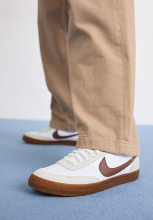 Persona che indossa pantaloni beige e sneakers bianche con il logo Nike marrone e suole in gomma, in piedi su un tappeto blu.
