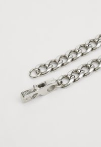 Zilveren metalen ketting met dikke schakels en een sluiting met het merk "GUESS." Gladde en glanzende textuur, met gegraveerde details op de sluiting.